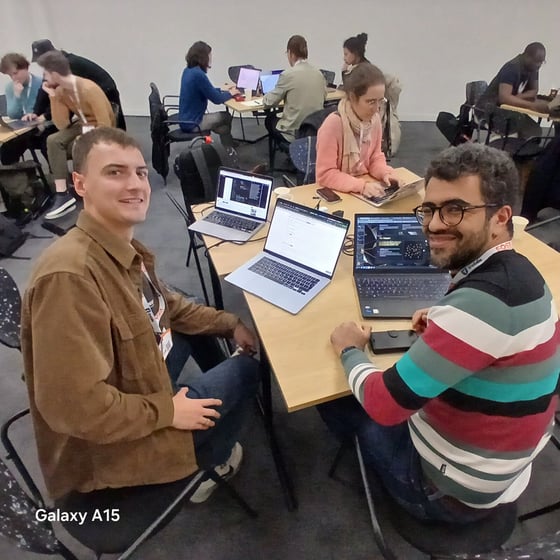 Two men smile directly at the camera while seated at a table with laptops during an indoor workshop or coding session, surrounded by several other participants working in the background.