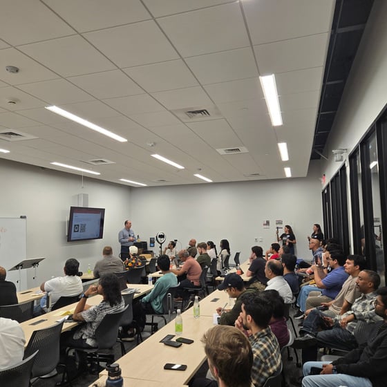 A photograph capturing a presentation or lecture taking place in a modern, brightly lit conference room filled with a large audience seated at tables. A male presenter stands at the front addressing the attendees, with content displayed on a wall-mounted monitor behind him.