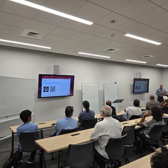 A photograph capturing an indoor training session or presentation, showing approximately a dozen attendees seated at desks facing a presenter and two wall-mounted screens displaying a webpage with a QR code.