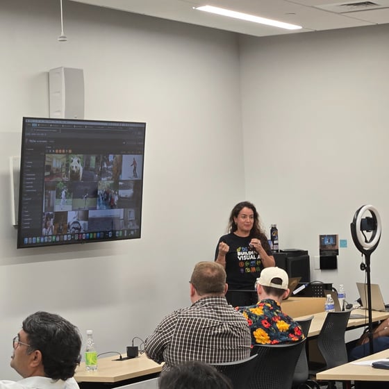 A woman delivers a presentation or lecture in a well-lit classroom, standing next to a large wall-mounted monitor displaying a media library interface, while several attendees listen from the foreground.