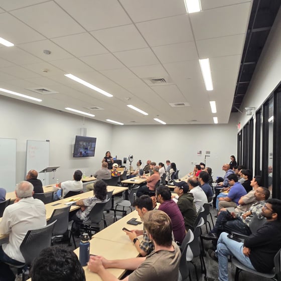 A photo capturing a large audience seated at tables in a modern, well-lit conference room, listening to a woman giving a presentation at the front of the room. A large monitor displaying a video feed and a whiteboard are visible behind the presenter.