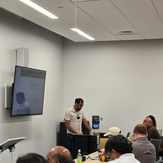 A photograph captures an indoor presentation setting where a man stands next to a large wall-mounted screen, addressing a group of seated attendees in a modern, brightly lit conference room. The screen displays text content and a strong reflection of the presenter's silhouette.