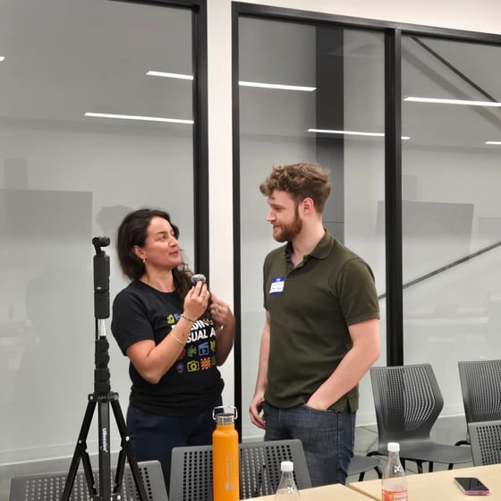 A candid photograph showing a woman holding a microphone and speaking to a man in a modern, brightly lit meeting room setting with glass partitions. A tripod and various water bottles are visible in the foreground.