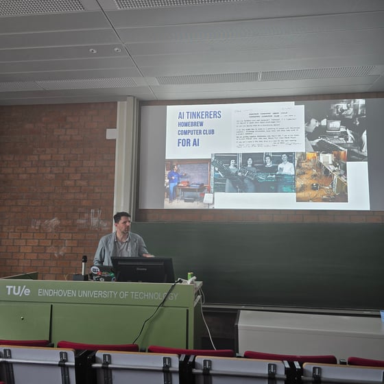 A man stands at a podium giving a presentation in a lecture hall at Eindhoven University of Technology, with a slide about the 'AI Tinkerers Homebrew Computer Club' projected behind him.