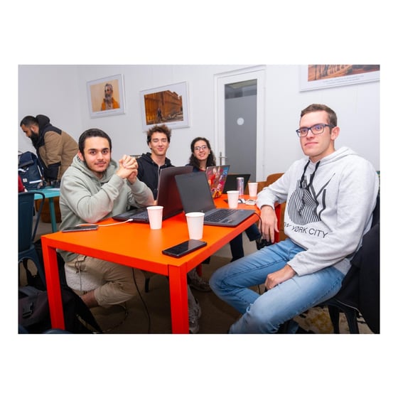 A candid photograph showing five young adults gathered around a bright orange table in an indoor setting, with four of them seated and working on laptops.