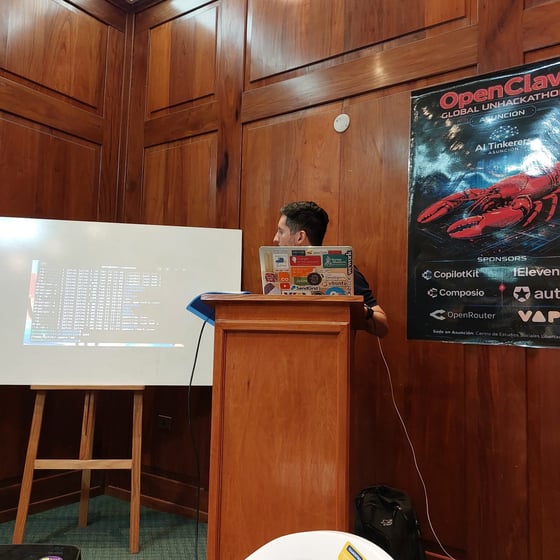 A man stands behind a wooden podium giving a presentation, with a terminal window projected onto a whiteboard and an event poster for a hackathon on the wood-paneled wall.