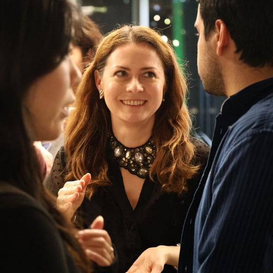 A close-up, candid photograph captures a smiling woman with long brown hair and a large, ornate jeweled necklace engaged in conversation between two other individuals, likely at an indoor social or networking event.
