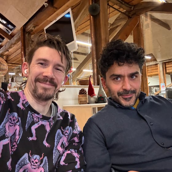 A close-up photo featuring two men smiling directly at the camera while sitting indoors in a rustic setting characterized by exposed wooden beams and warm lighting.