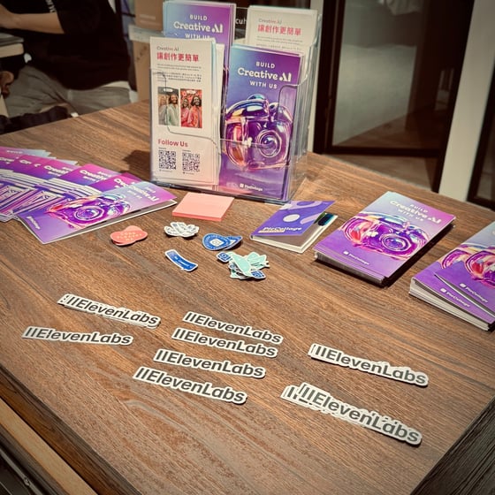 A close-up photograph of a wooden table displaying promotional materials, including flyers, notebooks, and stickers for companies like PicCollage and ElevenLabs, likely at a conference or event booth.