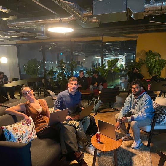 A candid photograph captures a group of six people working and socializing in a modern, dimly lit office lounge or co-working space at night, featuring industrial ceilings and large windows overlooking city lights. Three individuals in the foreground are smiling at the camera while using laptops.
