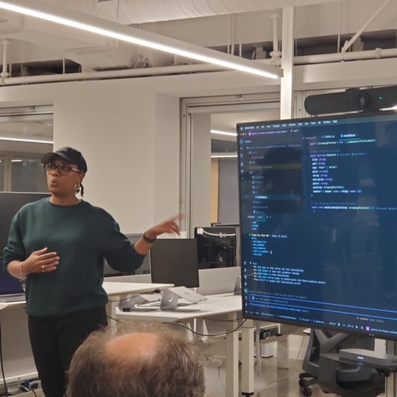 A woman wearing a dark green sweater and a baseball cap is giving a technical presentation indoors, gesturing toward a large monitor displaying lines of code in a dark mode environment. The top of an audience member's head is visible in the foreground.