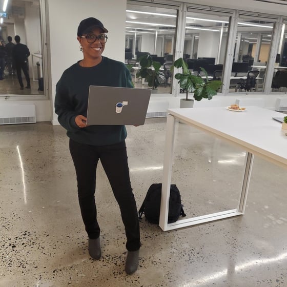 A smiling woman wearing a dark green sweater, black pants, and a baseball cap stands in a modern office space, holding a closed grey laptop. The setting features polished concrete floors, a large white table, and glass-walled offices in the background.