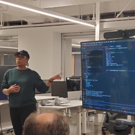 A woman wearing a dark green sweater and a baseball cap is giving a technical presentation in a modern office, pointing toward a large screen displaying lines of code.