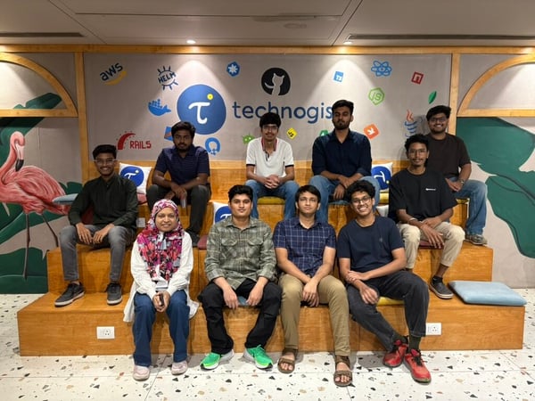 A group of ten young people are posing for a photo while sitting on wooden tiered seating in an indoor space decorated with tech logos.