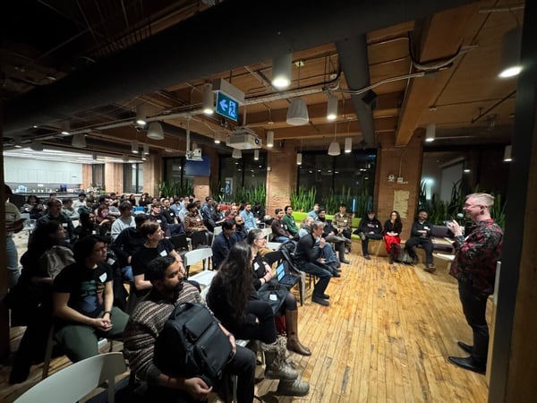 A man stands at the front of a room with wooden floors and exposed brick walls, addressing a seated audience during a presentation or event.