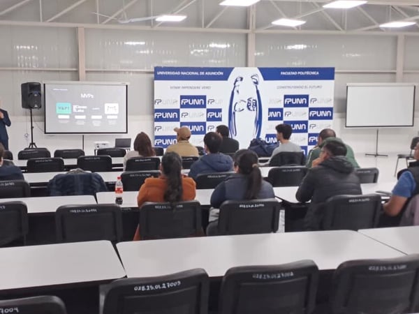 A photograph of a presentation being held in a university classroom, where a speaker in a suit stands next to a projector screen displaying a slide about 'MVPs' while an audience watches from their desks.