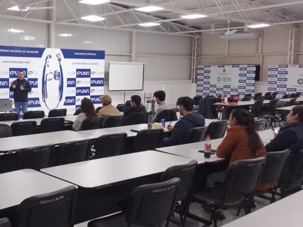 A man is giving a presentation in a university classroom, standing in front of a projector screen and a banner, while a small audience sits at desks listening.