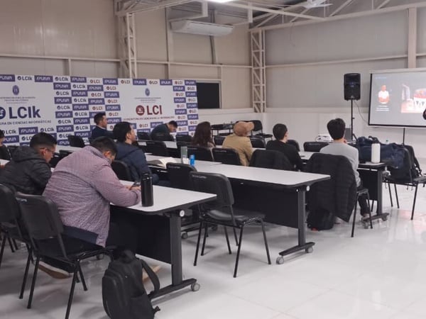 A photograph showing a presentation or lecture inside a university classroom, where a speaker stands at the front next to a projector screen and banners, while an audience sits at desks listening.