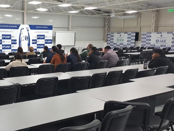 An indoor photograph of a lecture hall or classroom setup with rows of empty desks in the foreground and a small audience seated near the front facing a presentation screen and banners.