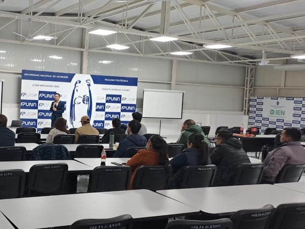An indoor photograph of a presentation or lecture taking place inside a hall, with a presenter standing in front of a blue and white backdrop and an audience seated at rows of desks.