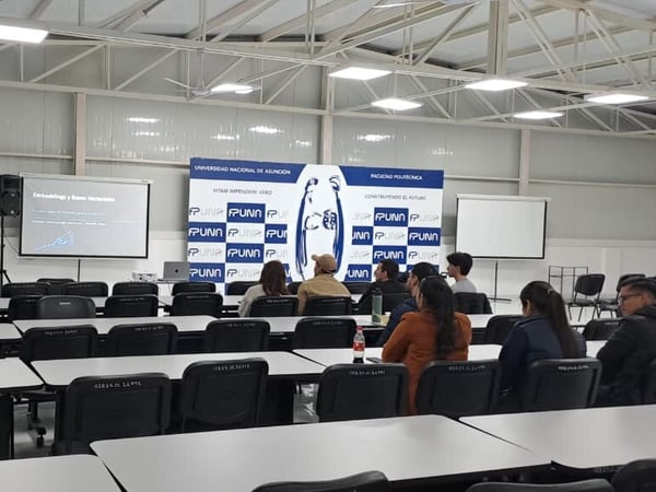 A presentation is being delivered in a large classroom or hall with rows of empty desks in the foreground and a small audience seated near the front screen.