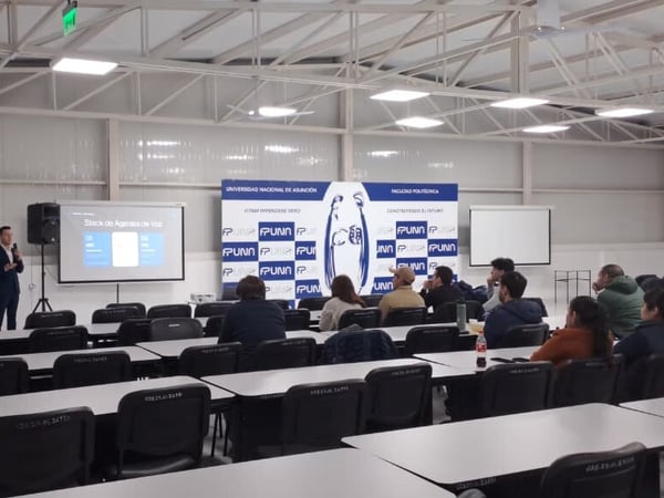 An indoor photograph showing a speaker giving a presentation titled 'Stack de Agentes de Voz' to an audience seated at tables in a large hall with FPUNA branding.