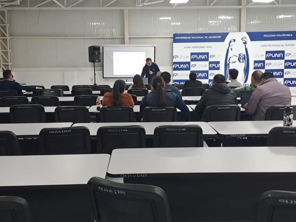 An indoor photograph of a university presentation or lecture hall, showing a presenter standing at the front near a projector screen with an audience seated at desks facing him.