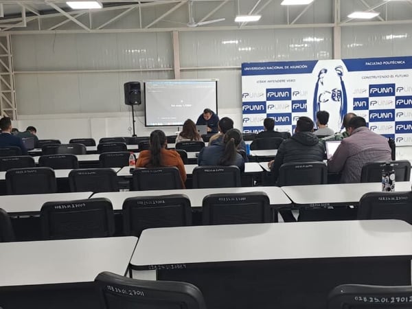 An indoor photo of a lecture hall or classroom at the Universidad Nacional de Asunción, showing rows of desks and chairs with several people seated and facing a presentation screen at the front.