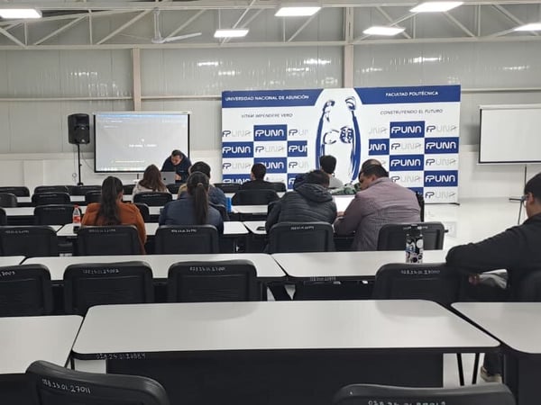 An indoor photograph of a university classroom or lecture hall at the Facultad Politécnica of the Universidad Nacional de Asunción, showing several rows of empty desks and a small audience facing a presenter at the front.