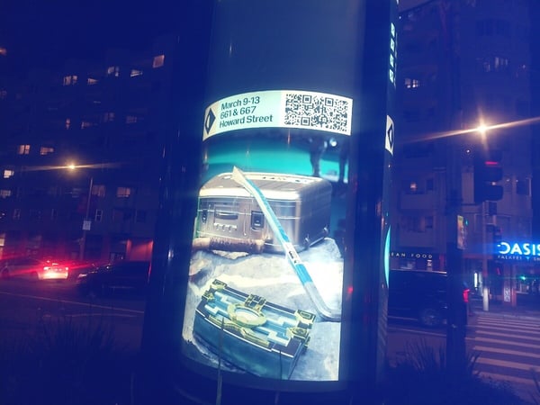 A night photograph of an illuminated advertising pillar on a city street, displaying an ad for an event with a QR code and an image of futuristic-looking chests and a tool.