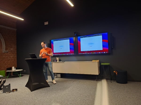 A man in an orange shirt is giving a presentation in a modern, dark-walled room, standing behind a black draped table with a laptop, while two large screens behind him display a loading browser page.