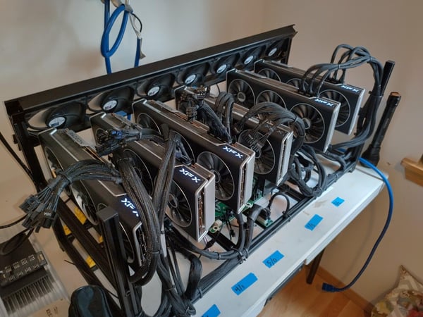 A photograph of a cryptocurrency mining rig setup, featuring multiple black Graphics Processing Units (GPUs) branded XFX and Radeon mounted in an open metal frame, resting on a white table.