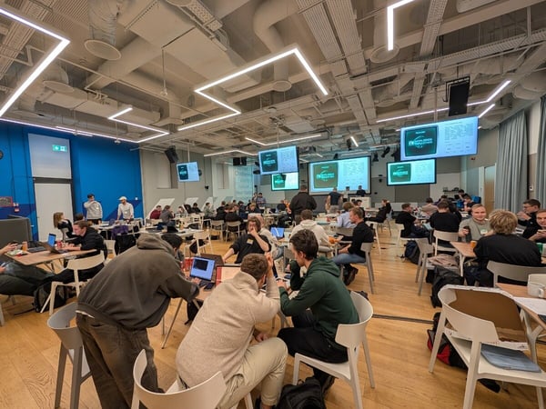A candid photograph captures a busy indoor event, likely a hackathon or workshop, where numerous participants are seated at tables working on laptops beneath modern, angular ceiling lights. Large projection screens are visible displaying content related to the activity.
