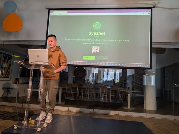 A young man, dressed in a brown hoodie and cargo pants, stands at a clear acrylic podium presenting from a laptop, with a large screen behind him displaying the homepage for a service called 'Syuzhet'.