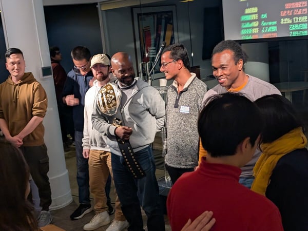 A photograph captures a group of diverse individuals gathered indoors at an event, focusing on a man in the center who is proudly holding a large championship belt.