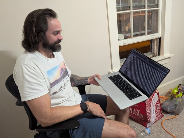 A man with long hair and a beard sits in an office chair indoors, holding a laptop open to a screen displaying lines of code or logs, with a cluttered background including a window and a red shopping bag.