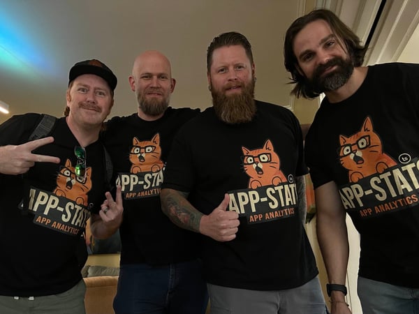 Four men wearing identical black t-shirts featuring an orange cat graphic and the text 'APP-STATS APP ANALYTICS' pose together for a group photo in a dimly lit indoor setting.