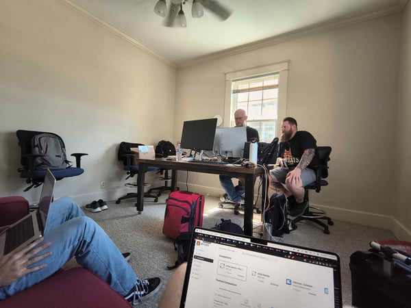 A candid photograph showing three individuals working on computers in a brightly lit, sparsely furnished indoor office or temporary workspace, with the view partially obscured by the laptop screen of the person in the foreground.
