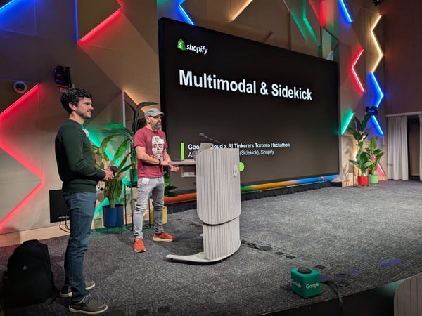 Two men are standing on a stage in front of a large projection screen displaying a presentation titled "Multimodal & Sidekick" during a Google Cloud x AI Tinkerers Toronto Hackathon event hosted by Shopify. The stage features a modern, geometrically paneled background illuminated with colorful neon lights.