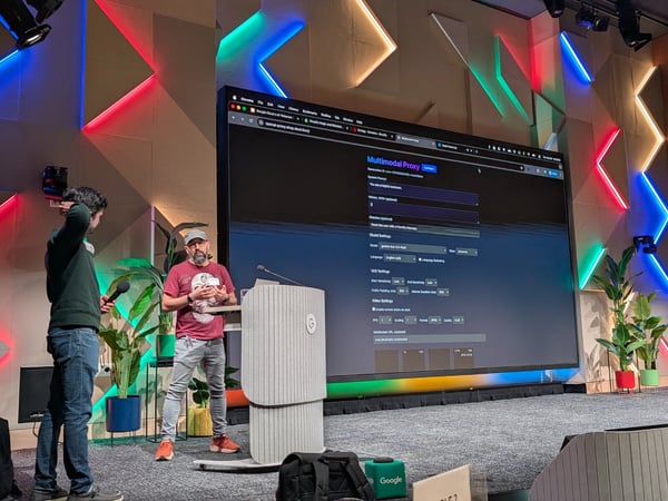 A photograph capturing a technical presentation on a stage, where two men stand in front of a very large screen displaying a web interface for a "Multimodal Proxy." The stage features a modern, geometric background illuminated by red, green, and blue neon lights.