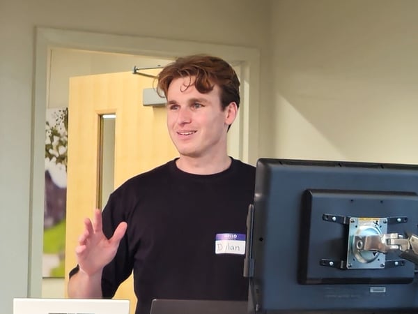 A candid indoor photo showing a young man, wearing a name tag that reads "Dylan," smiling and gesturing while standing next to a computer monitor, likely giving a presentation.