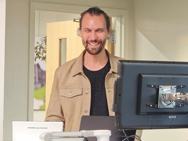 A man with long dark hair and a beard, wearing a beige jacket, stands smiling broadly behind a workstation setup featuring a monitor and laptop in an indoor office environment.