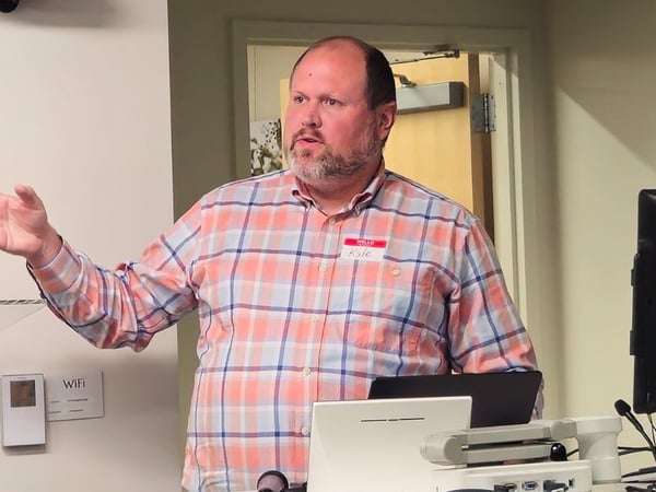 A man wearing a salmon and blue plaid shirt and a name tag is giving a presentation or talk in an indoor setting, gesturing with his right hand near a clear acrylic podium and computer equipment.