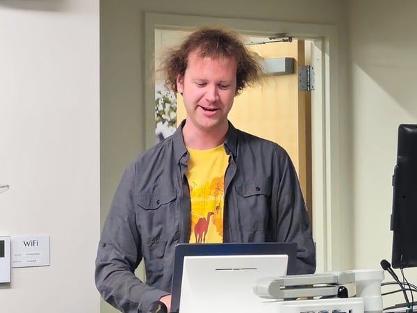A man with messy hair, wearing a gray shirt over a yellow graphic t-shirt, stands in an indoor setting, interacting with a laptop and a monitor on a stand, likely preparing for or giving a presentation.