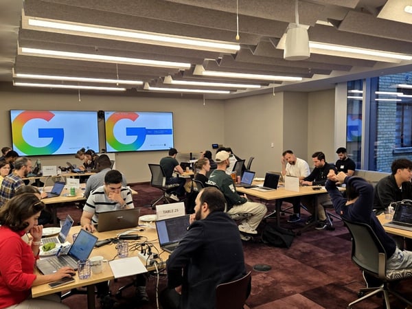 A photograph capturing a large group of people working intently on laptops at tables in a modern office or event space, likely during a coding session or hackathon. Two large screens on the back wall prominently display the colorful Google 'G' logo.