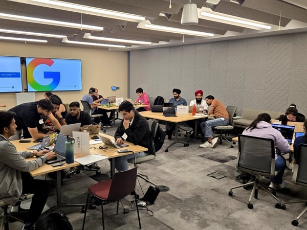 A photograph showing a group of approximately seventeen people working on laptops at tables in a modern, well-lit collaborative workspace or classroom. Two large screens on the front wall prominently display the Google logo and a search interface.