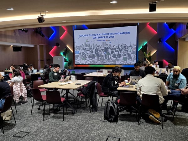 A photograph capturing a busy indoor hackathon or coding event, showing numerous participants seated at tables working intently on laptops. A large screen in the background displays the event title and date.
