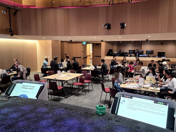 A photograph captures a large group of people seated at tables working on laptops in a modern auditorium or event space, likely participating in a hackathon or workshop. Two digital displays are visible in the foreground, one displaying a presentation slide about 'Agentic AI'.