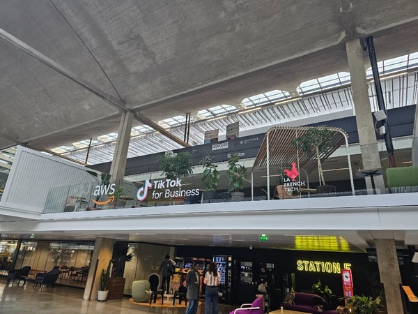 A photograph showing the modern, multi-level interior of a large building, likely a startup campus or innovation hub, featuring prominent corporate logos like AWS, TikTok for Business, and La French Tech on the upper level.