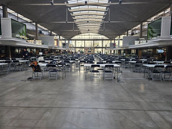 A wide-angle photograph of a vast, high-ceilinged indoor hall, likely a convention center or former industrial space, set up with hundreds of rows of empty white folding tables and black chairs, suggesting preparation for a large event.